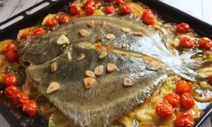 Rodaballo al horno con tomates cherry y láminas de ajo, servido sobre una cama de cebolla caramelizada en una bandeja de horno.
