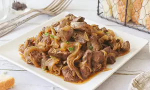 Sabrosos higaditos de pollo encebollados, servidos en un plato blanco, con pan y tenedores al fondo.