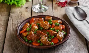 Un plato de carne en salsa con guisantes y zanahorias, servido en una cazuela de barro sobre una mesa de madera rústica.