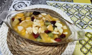 Deliciosa macedonia de frutas en un cuenco de cristal, con trozos de kiwi, plátano, manzana y frutos rojos en almíbar.