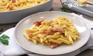 Plato de macarrones a la carbonara cremosos con trozos de panceta. En el fondo, un recipiente con más pasta y un rallador de queso.