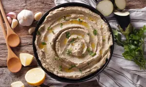 Un cremoso hummus de berenjena, adornado con aceite de oliva y perejil, junto a limones, ajos y cucharas de madera.