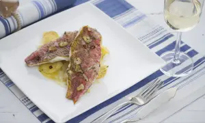 Dos deliciosos salmonetes al horno, con patatas y ajo, servidos en un plato blanco junto a una copa de vino blanco.