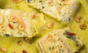 Merluza al pil pil, un plato tradicional con trozos de pescado blanco en una salsa cremosa, adornado con láminas de ajo dorado y guindilla.