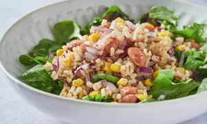 Ensalada de arroz con salchicha, maíz, cebolla morada y frijoles, servida sobre hojas verdes frescas.