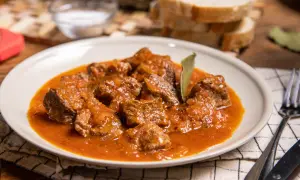 Sabrosa carne con tomate, guisada lentamente hasta quedar tierna. Servida en un plato blanco con pan y cubiertos.