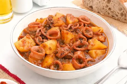 Un tazón de patatas con calamares en salsa, acompañado de pan rústico y una pizca de pimentón.