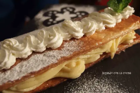 Delicioso milhojas de crema, con hojaldre crujiente y relleno cremoso, decorado con nata montada y una hoja de menta.