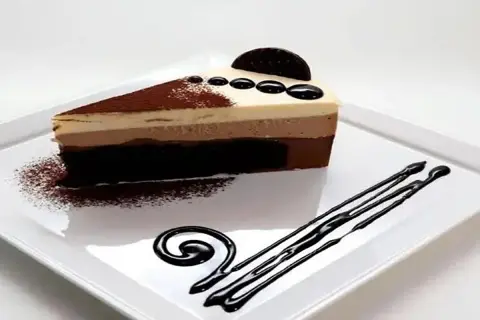 Deliciosa tarta de 3 chocolates, con capas de mousse y bizcocho, decorada con cacao y un toque de chocolate líquido.