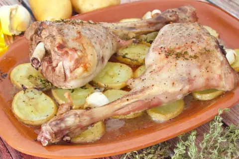 Dos jugosas piernas de cabrito al horno, tiernas y doradas, reposan sobre patatas asadas y dientes de ajo.