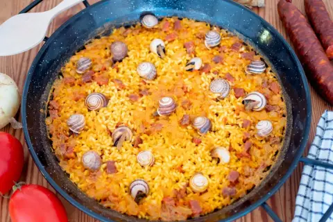 Arroz con caracoles en paellera, con trozos de chorizo y decorado con caracoles. Ingredientes frescos alrededor.