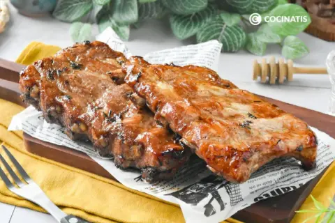 Costillas a la miel, jugosas y glaseadas, servidas sobre papel de periódico y tabla de madera.