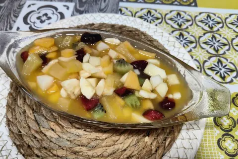 Deliciosa macedonia de frutas en un cuenco de cristal, con trozos de kiwi, plátano, manzana y frutos rojos en almíbar.