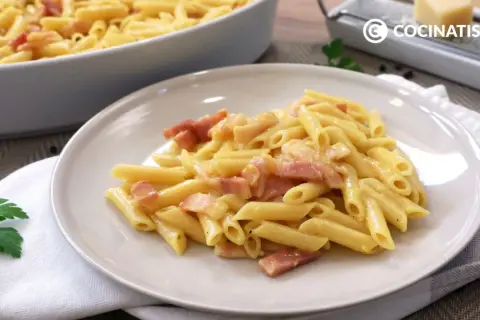 Plato de macarrones a la carbonara cremosos con trozos de panceta. En el fondo, un recipiente con más pasta y un rallador de queso.