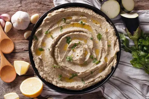 Un cremoso hummus de berenjena, adornado con aceite de oliva y perejil, junto a limones, ajos y cucharas de madera.