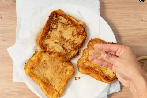 Torrijas de horchata doradas y crujientes, listas para disfrutar. Una mano espolvorea azúcar y canela sobre una de ellas.