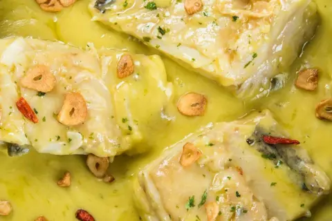 Merluza al pil pil, un plato tradicional con trozos de pescado blanco en una salsa cremosa, adornado con láminas de ajo dorado y guindilla.