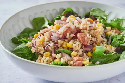 Ensalada de arroz con salchicha, maíz, cebolla morada y frijoles, servida sobre hojas verdes frescas.