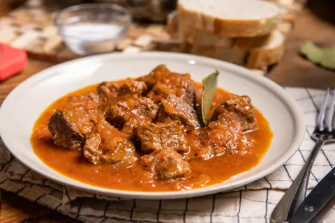 Sabrosa carne con tomate, guisada lentamente hasta quedar tierna. Servida en un plato blanco con pan y cubiertos.
