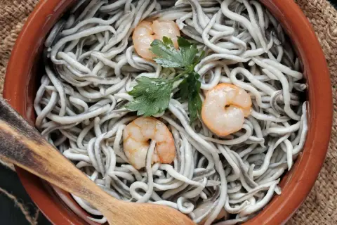 Gulas al ajillo servidas en cazuela de barro, con gambas y perejil. Una cuchara de madera lista para servir.