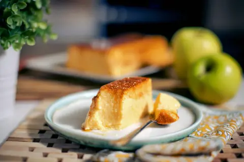 Un delicioso flan de manzana, con una porción servida en plato, junto a manzanas verdes y una planta.