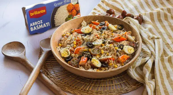 Delicioso arroz con atún, huevo duro, aceitunas y maíz en un bol de madera. Acompañado de cucharas de madera y una caja de arroz basmati.