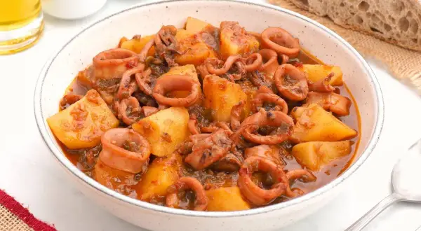 Un tazón de patatas con calamares en salsa, acompañado de pan rústico y una pizca de pimentón.