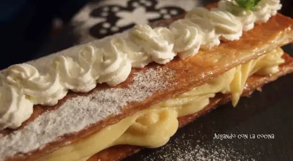 Delicioso milhojas de crema, con hojaldre crujiente y relleno cremoso, decorado con nata montada y una hoja de menta.