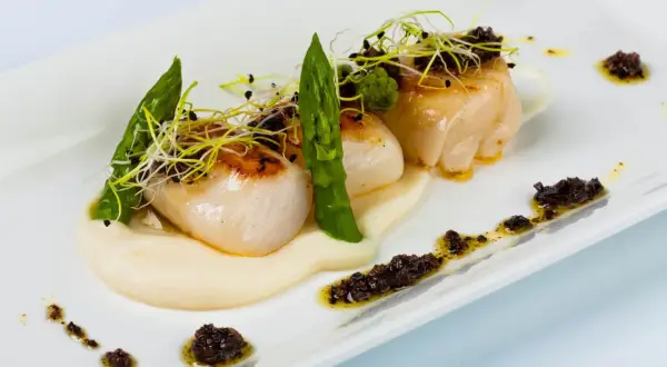 Deliciosas vieiras a la plancha sobre puré cremoso, adornadas con espárragos tiernos y brotes frescos. Un plato elegante y sabroso.
