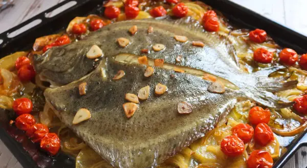 Rodaballo al horno con tomates cherry y láminas de ajo, servido sobre una cama de cebolla caramelizada en una bandeja de horno.