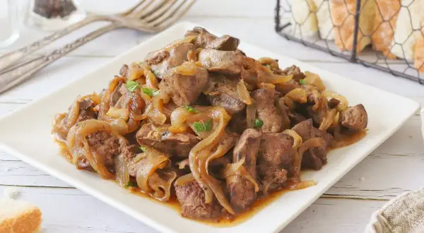 Sabrosos higaditos de pollo encebollados, servidos en un plato blanco, con pan y tenedores al fondo.
