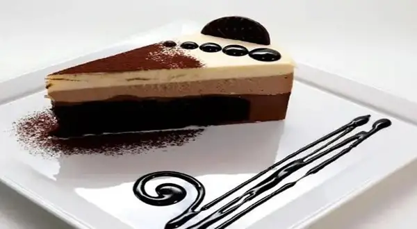 Deliciosa tarta de 3 chocolates, con capas de mousse y bizcocho, decorada con cacao y un toque de chocolate líquido.
