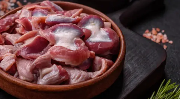 Un cuenco de madera lleno de mollejas de pollo crudas, listas para ser cocinadas. Sal rosada y romero al lado.