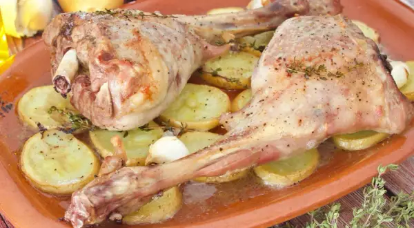 Dos jugosas piernas de cabrito al horno, tiernas y doradas, reposan sobre patatas asadas y dientes de ajo.