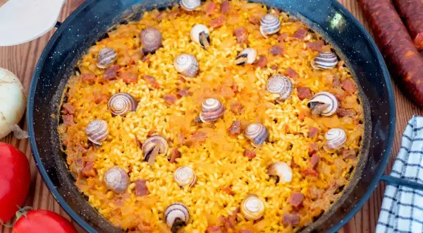 Arroz con caracoles en paellera, con trozos de chorizo y decorado con caracoles. Ingredientes frescos alrededor.