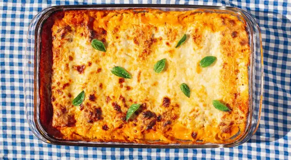 Bandeja de canelones de espinacas gratinados, decorados con hojas de albahaca fresca, sobre un mantel de cuadros azules y blancos.