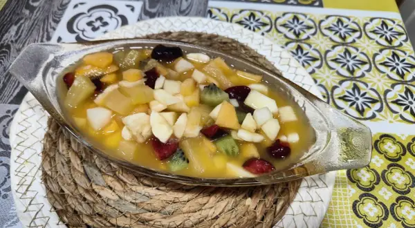 Deliciosa macedonia de frutas en un cuenco de cristal, con trozos de kiwi, plátano, manzana y frutos rojos en almíbar.
