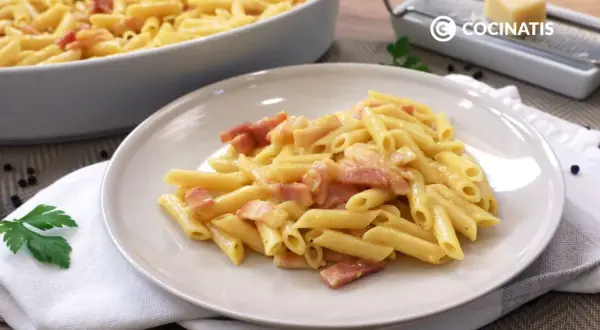 Plato de macarrones a la carbonara cremosos con trozos de panceta. En el fondo, un recipiente con más pasta y un rallador de queso.