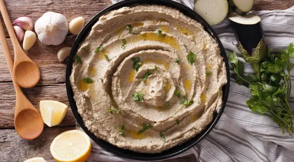 Un cremoso hummus de berenjena, adornado con aceite de oliva y perejil, junto a limones, ajos y cucharas de madera.