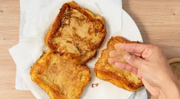 Torrijas de horchata doradas y crujientes, listas para disfrutar. Una mano espolvorea azúcar y canela sobre una de ellas.