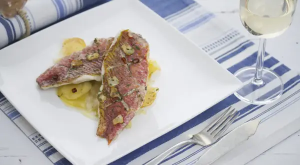 Dos deliciosos salmonetes al horno, con patatas y ajo, servidos en un plato blanco junto a una copa de vino blanco.