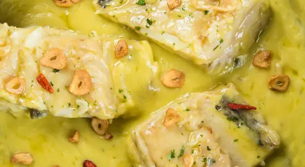 Merluza al pil pil, un plato tradicional con trozos de pescado blanco en una salsa cremosa, adornado con láminas de ajo dorado y guindilla.