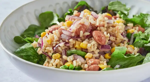 Ensalada de arroz con salchicha, maíz, cebolla morada y frijoles, servida sobre hojas verdes frescas.