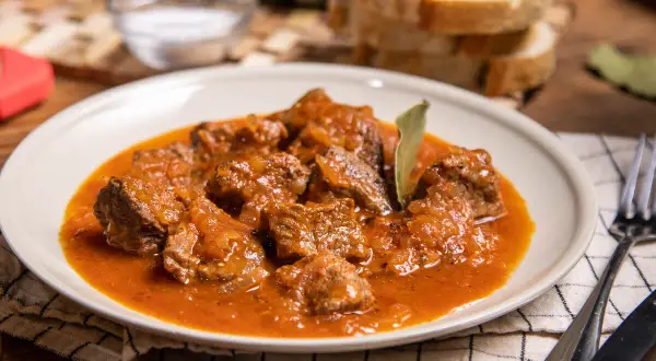 Sabrosa carne con tomate, guisada lentamente hasta quedar tierna. Servida en un plato blanco con pan y cubiertos.