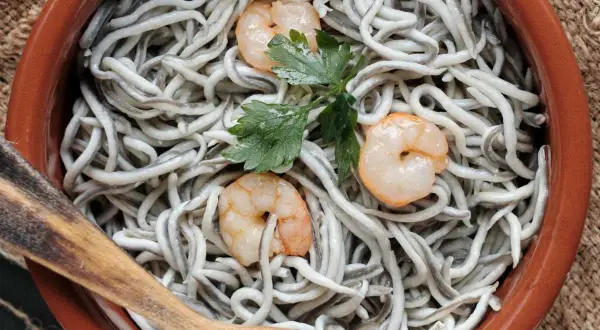 Gulas al ajillo servidas en cazuela de barro, con gambas y perejil. Una cuchara de madera lista para servir.