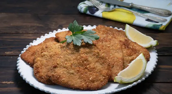 Milanesa de ternera dorada y crujiente, servida en plato blanco con gajos de limón y perejil.