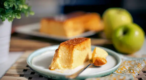 Un delicioso flan de manzana, con una porción servida en plato, junto a manzanas verdes y una planta.