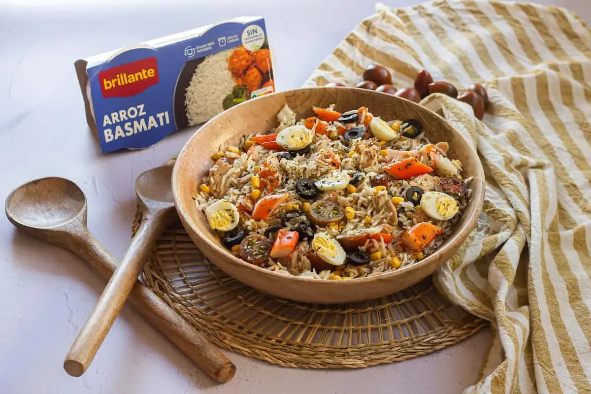 Delicioso arroz con atún, huevo duro, aceitunas y maíz en un bol de madera. Acompañado de cucharas de madera y una caja de arroz basmati.