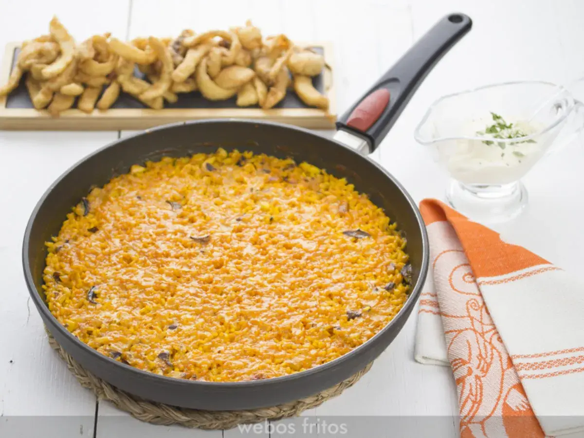 Arroz con sepia en paellera, acompañado de patatas fritas y una salsa cremosa.