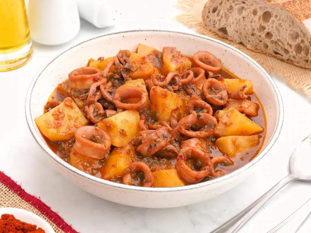 Un tazón de patatas con calamares en salsa, acompañado de pan rústico y una pizca de pimentón.